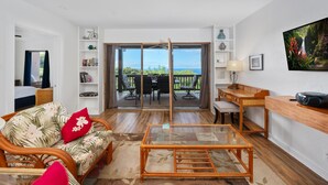 A living room with a sofa, a glass coffee table, a dining area with a table and chairs, and a balcony with a view. - Big Island Kona Westwind 12 2 Bedroom Condo (Kailua-Kona)