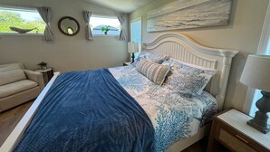 Un dormitorio con cama, mesita de noche, lámpara, ventana con cortinas, espejo en la pared y un cuadro.