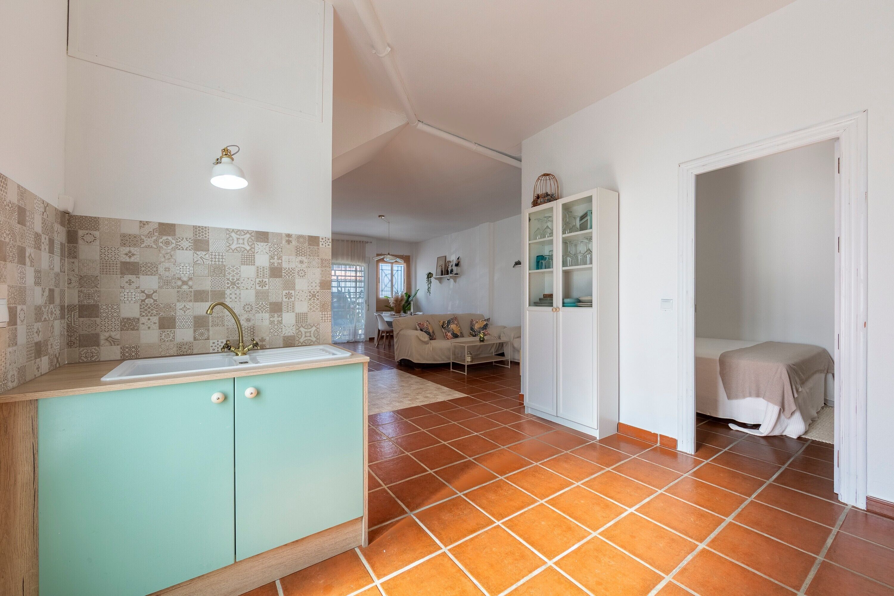 Een keuken met een spoelbak, kastjes en een decoratieve achterwand. Er is een eethoek met een tafel en stoelen, en een woonkamer met een bank.