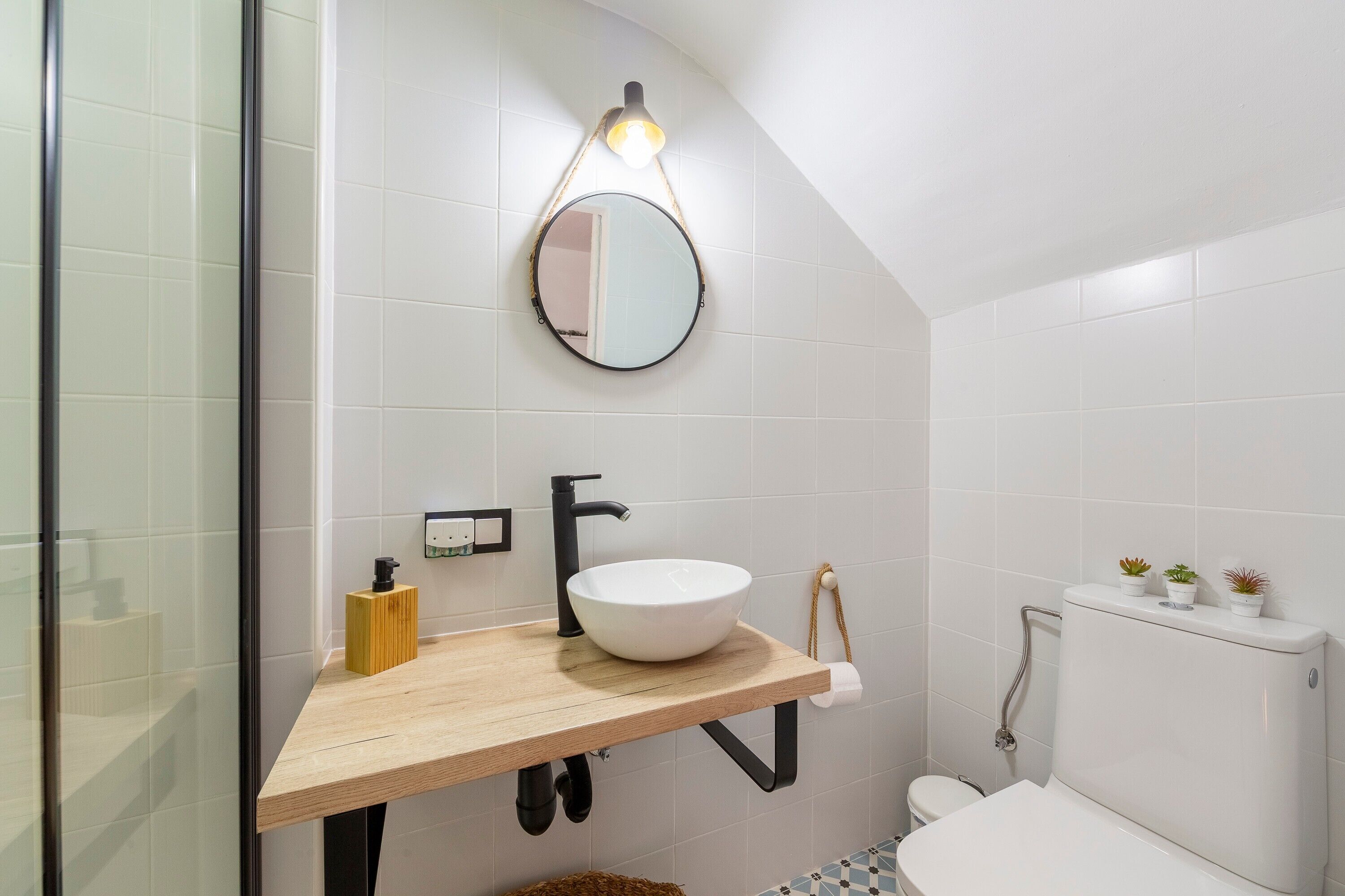 Un baño con tocador de madera, espejo redondo, lavamanos blanco, inodoro y toallero.