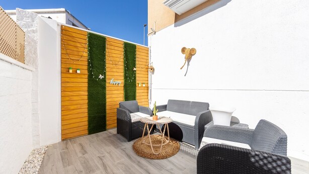 Un área de estar al aire libre con mobiliario de mimbre y una mesa pequeña, una valla de madera con follaje y un adorno de Mickey Mouse colgado en la pared.