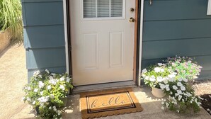 Une porte d’entrée ornée d’une guirlande, surmontée d’un paillasson et flanquée de deux pots de fleurs.