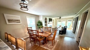 Un coin repas avec une table et des chaises en bois, un ventilateur de plafond et une vue sur le salon, où se trouvent un canapé et une plante.