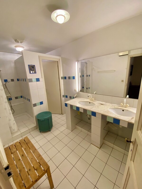 A bathroom with white tiled floors, a wooden bench, a green trash bin, and a double sink with a mirror above.
