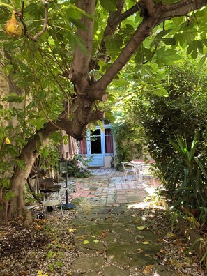 A garden pathway leading to a house with a blue door, surrounded by greenery and a rustic metal chair.