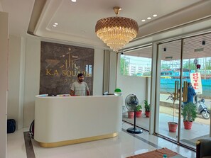 A hotel reception area with a modern chandelier, a reception desk, a wall with the hotel's name, and a view of the street outside.