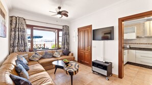 Sala de estar con un sofá de cuero, una mesa de centro, televisor y una cocina visible a través de una puerta abierta.