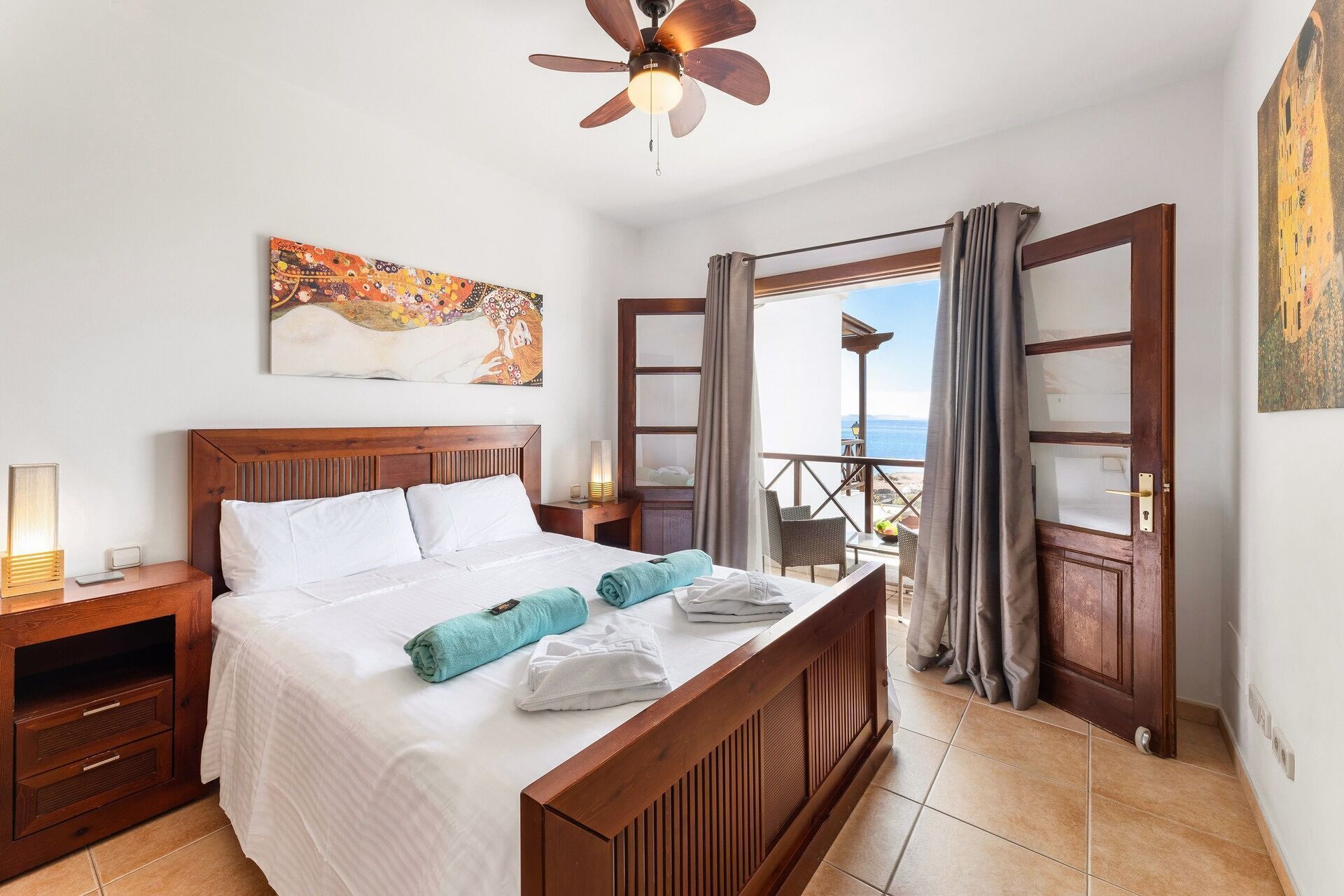 A hotel room with a wooden bed, a ceiling fan, a painting on the wall, and a balcony with a view of the sea.