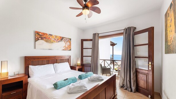 Habitación de hotel con una cama de madera, ventilador de techo, un cuadro en la pared y un balcón con vista al mar.
