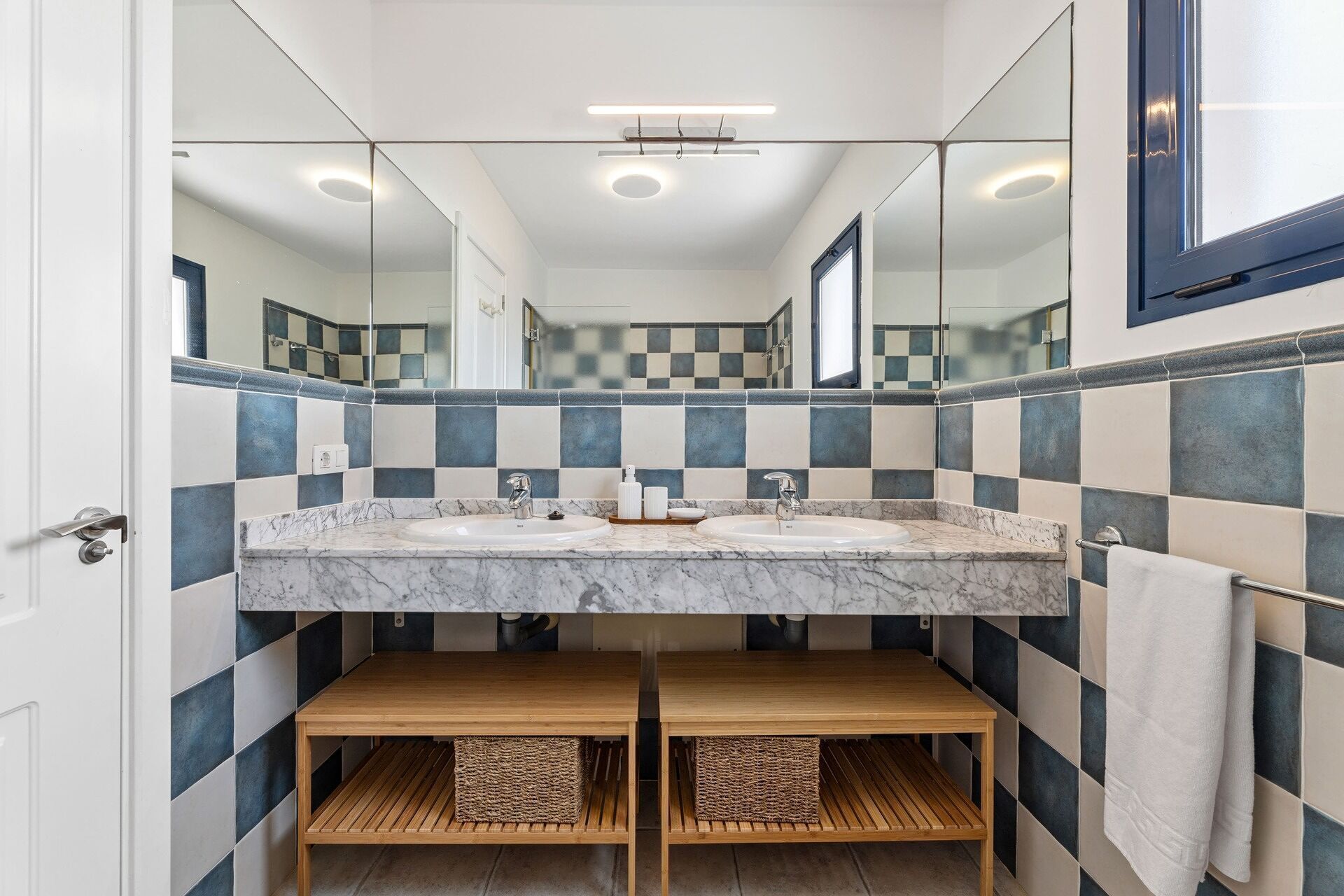 A bathroom with a checkered tile backsplash, two sinks with marble countertops, wooden benches, and a large mirror.