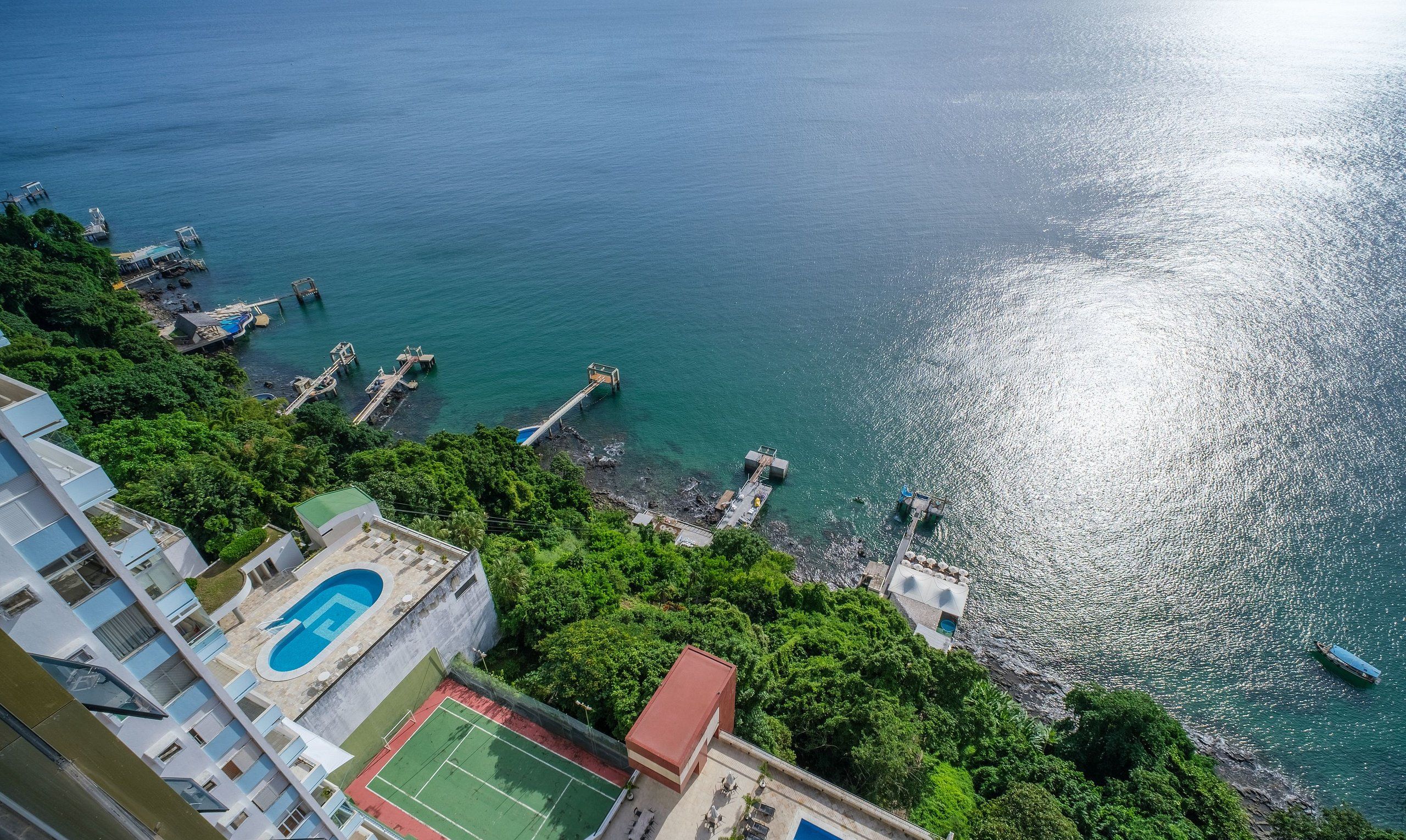 Vue aérienne d’une zone côtière avec une piscine, un court de tennis et une jetée qui s’avance dans l’eau.