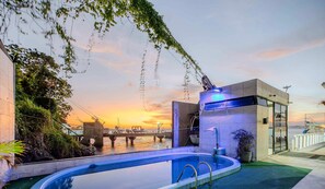 Une piscine avec vue sur un coucher de soleil se reflétant sur l’eau, un bâtiment moderne et un pont en arrière-plan.