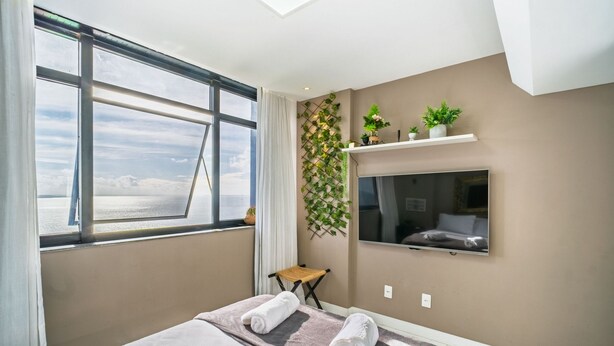 Une chambre d’hôtel moderne dotée d’une grande fenêtre, d’un lit avec des serviettes, d’un téléviseur fixé au mur et d’une étagère à plantes fixée au mur.