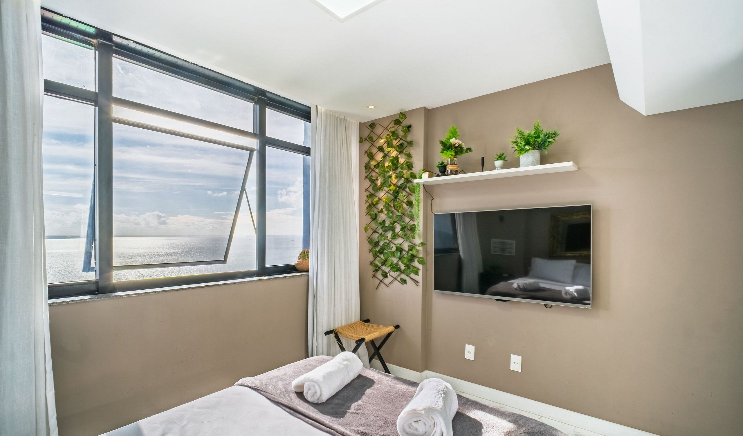 A hotel room with a large window, a bed with towels, a mounted TV, and a wall-mounted shelf with plants.