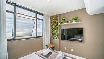 A hotel room with a large window, a bed with towels, a mounted TV, and a wall-mounted shelf with plants.