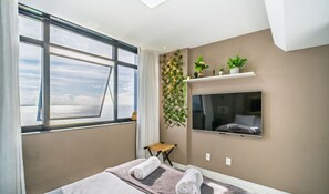 Habitació d'hotel amb una finestra gran, un llit amb tovalloles, un televisor fixat a la paret i una estant amb plantes.