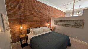 Ein Schlafzimmer mit Backsteinwand, einem Bett mit dunkelgrüner Bettdecke, zwei Nachttischlampen und einem Deckenventilator.