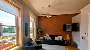 Ein Wohnzimmer mit Backsteinwand, Deckenventilator, Wand-Fernseher und großem Fenster mit Blick auf eine Baustelle.