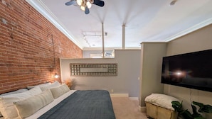 Ein Schlafzimmer mit Backsteinwand, einem Bett mit dunkler Bettdecke, einem weißen Nachttisch, einem Fernseher und einem Deckenventilator.
