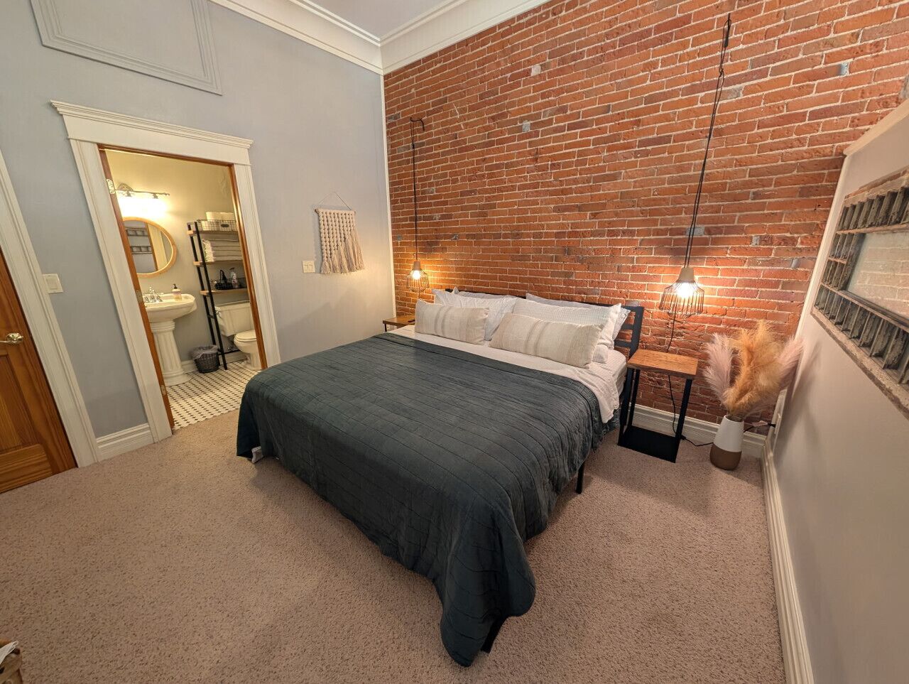Une chambre avec un mur en briques, un lit recouvert d’une couvre-lit vert foncé, une table de chevet avec une lampe et une petite table sur laquelle est posé un vase rempli de fleurs séchées.