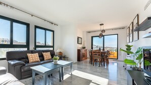 Un salon spacieux comprenant un canapé, une table basse et un coin repas. De grandes fenêtres offrent une vue sur la ville.