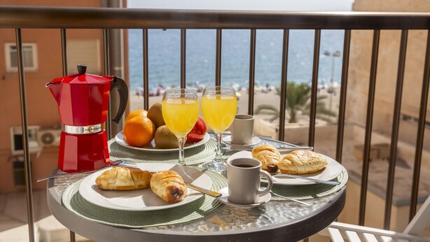 Une table de balcon avec un assortiment de produits pour le petit-déjeuner, notamment des croissants, du jus, du café et une cafetière rouge.
