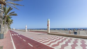 Une promenade en bord de mer bordée de palmiers, avec un chemin dallé aux motifs colorés et des parasols de plage visibles au loin.