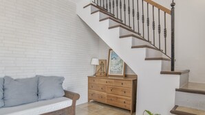 Una escalera con barandilla de madera y balaustradas de metal, un banco de madera con cojines, un aparador de madera con cajones y una planta en maceta.