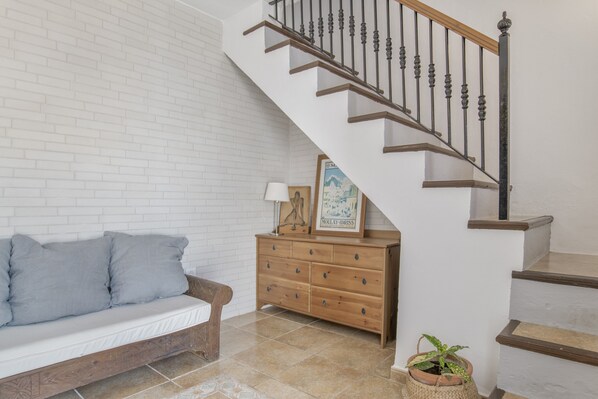 Una escalera con barandilla de madera y balaustradas de metal, un banco de madera con cojines, un aparador de madera con cajones y una planta en maceta.