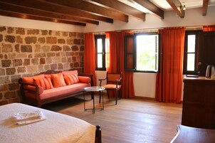 A room with a stone wall, wooden floor, a bed, a sofa, a chair, a small table, and a desk with a lamp.