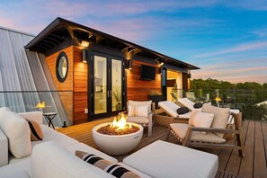 A modern outdoor deck with a fire pit, lounge chairs, and a glass balustrade overlooking a landscape.