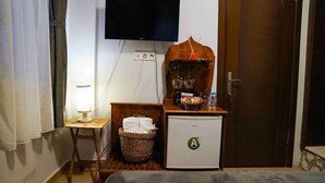 Ein kleines Hotelzimmer mit einem Bett, einem Tisch mit Lampe, einem Kühlschrank, einem an der Wand montierten Fernseher und einer Holzkommode mit Küchenutensilien.