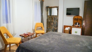 Ein Hotelzimmer mit einem Bett, zwei gelben Stühlen, einem kleinen Tisch mit Büchern, einem an der Wand befestigten Fernseher und einer Holzkabine mit einem Korb.
