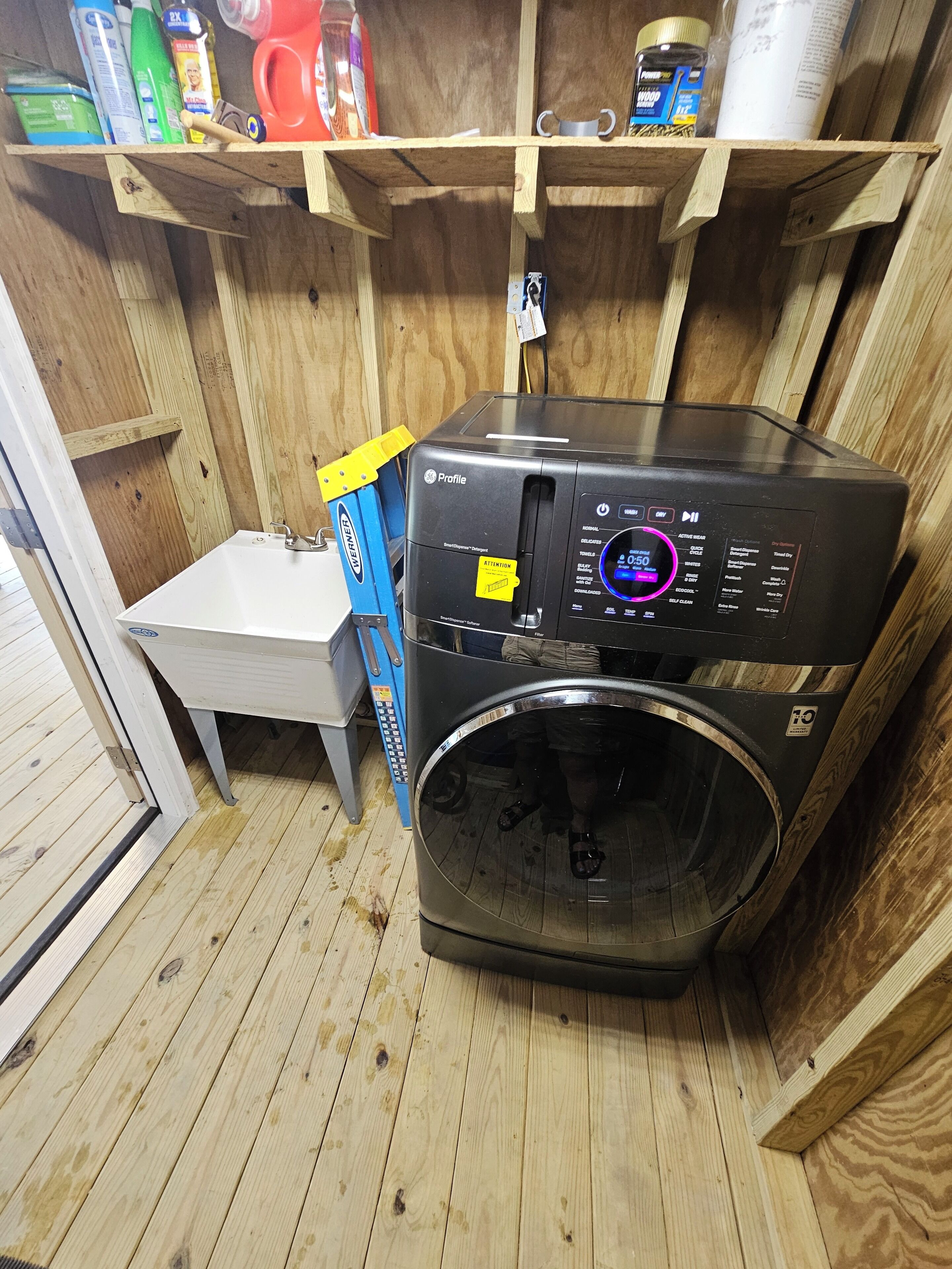 A compact laundry room with a front-loading washer and dryer, wooden shelving above, and a small table with a chair.