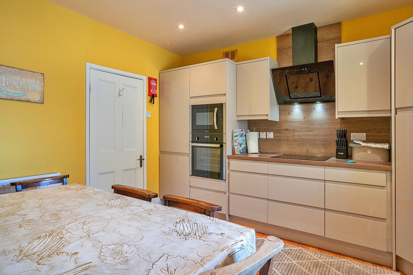 A compact kitchen with a microwave, oven, and a small countertop. There is a bed with a patterned bedspread and a wooden headboard. The room has yellow walls and a white door.