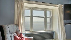 A room with a large window, a grey armchair, a red pillow, a black radiator, and a flat-screen TV mounted on the wall. - Beau View at Gadlys House (Beaumaris)