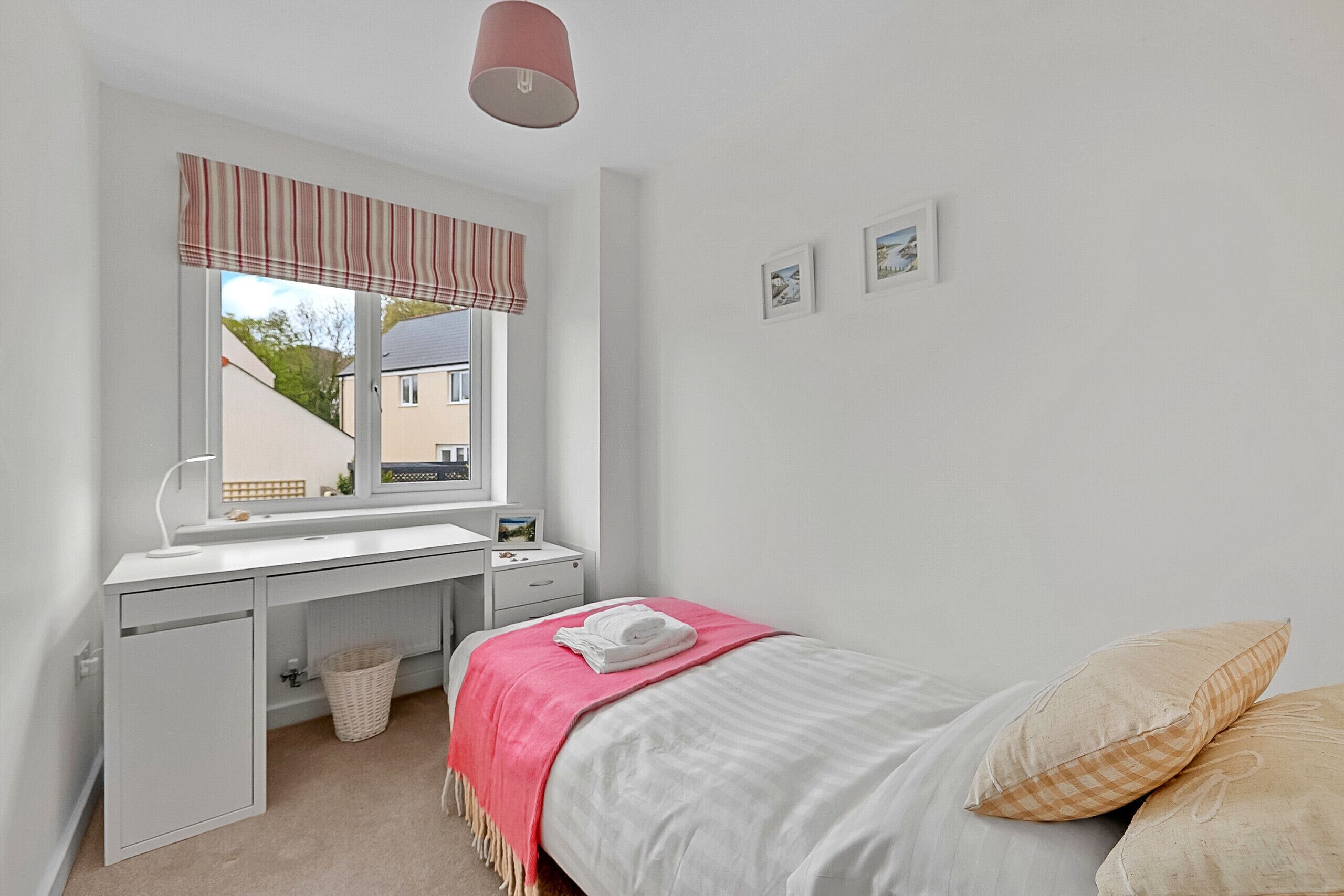 Ein Schlafzimmer mit Bett, Schreibtisch, Fenster mit Blick auf ein Haus und Wand mit gerahmten Bildern.