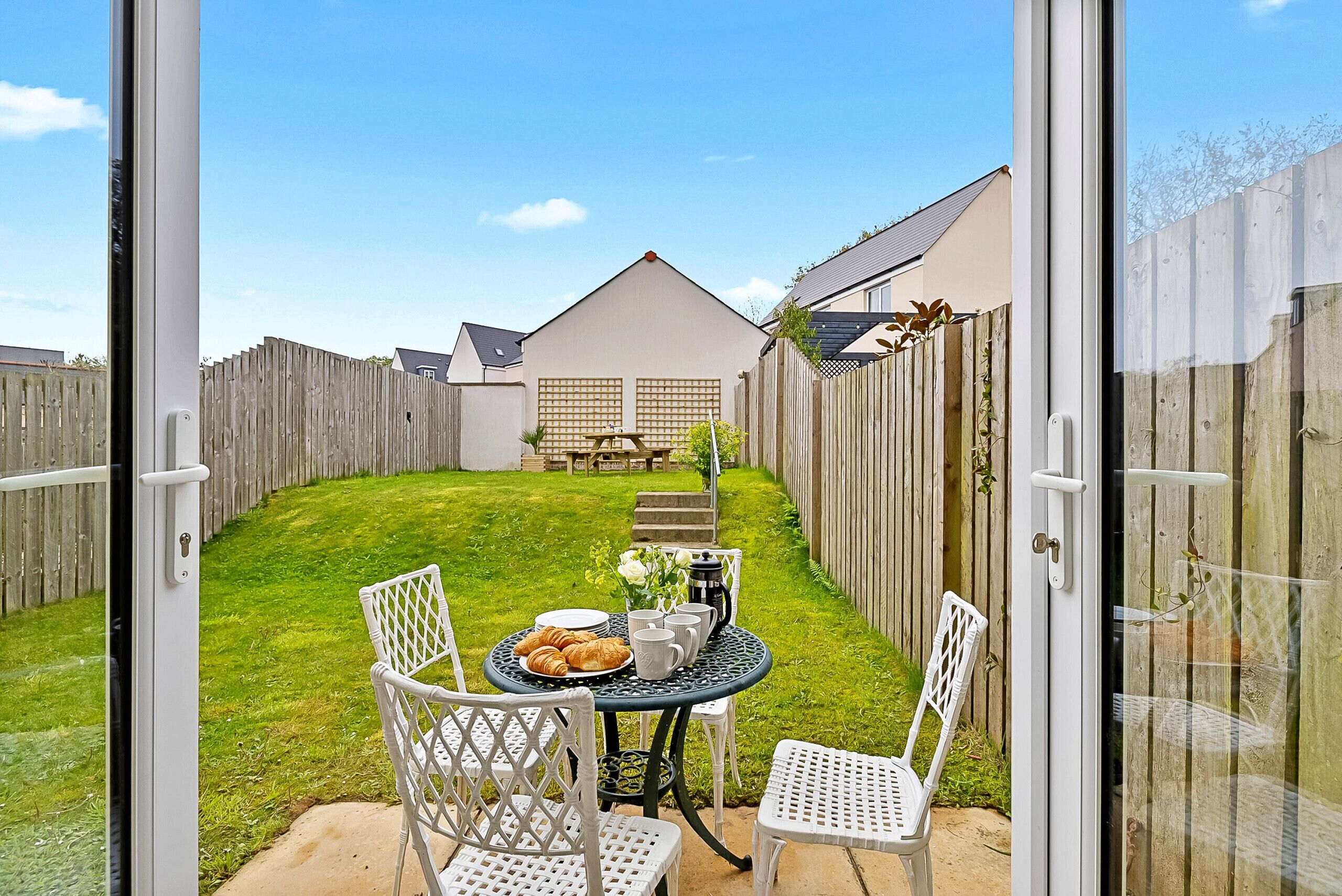 Une porte-fenêtre coulissante donne sur un jardin avec une table dressée pour le petit-déjeuner, des chaises blanches et une clôture en bois.