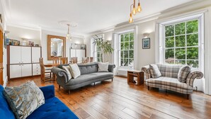 A spacious living room with hardwood flooring, a grey sofa, a blue sofa, and a plaid sofa. There are large windows with a view of greenery outside. - Palace Gate, Hampton Court (East Molesey)