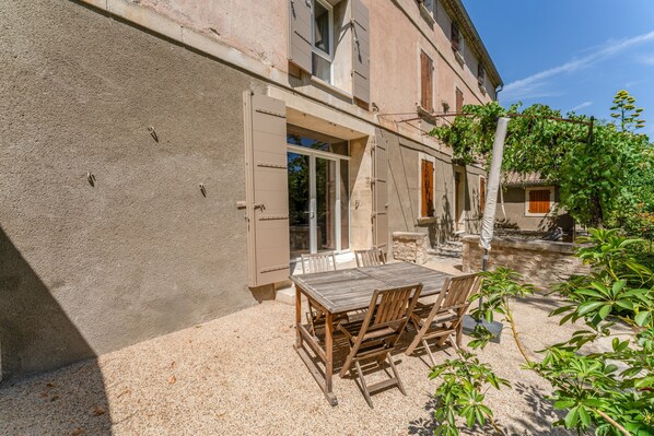 A patio with a wooden table and chairs, a stone wall, and a building with a window and door.