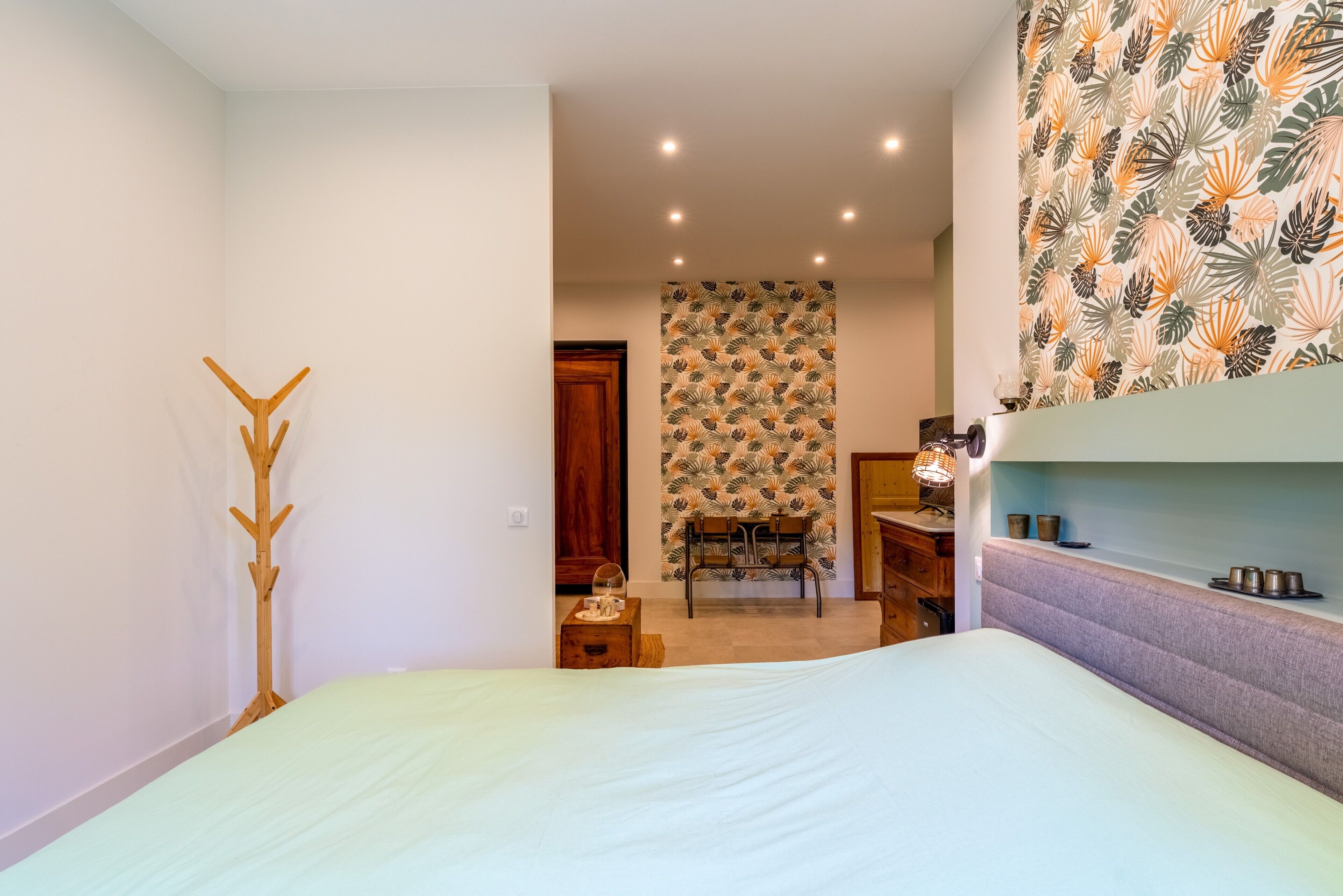 A bedroom with a bed, a coat rack, a patterned wall, a chair, and a shelf with items.