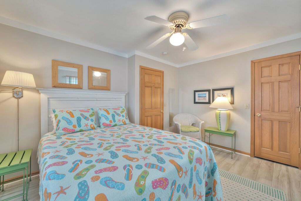 Un dormitorio con cama, ventilador de techo, dos cuadros enmarcados, una silla, una mesita de noche y dos puertas.
