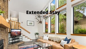 A modern living room with a fireplace, a large window, and a staircase leading to an upper level. - Peaceful Pines | Home Gym & Office, Near Trails (Fort Collins)