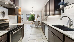 A modern kitchen with dark cabinetry, a stainless steel range hood, a double sink, and a dining area with a chandelier. - Peaceful Pines | Home Gym & Office, Near Trails (Fort Collins)
