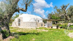 Una casa blanca de forma abovedada con muros y puerta de piedra, rodeada de vegetación y árboles.