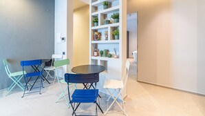 Una habitación con una mesa redonda, cuatro sillas, una estantería blanca con plantas y una pared gris.