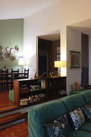 Sala de estar con sofá verde, una estantería, un comedor con sillas y una pared decorada con platos y marcos con fotografías.