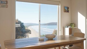 A dining area with a wooden table and chairs, a bowl, and a view of a beach and ocean through a large window.