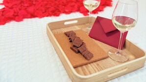 Un lit recouvert d’une courtepointe ornée de pétales de rose rouge, un plateau avec des morceaux de chocolat et deux verres de vin blanc, et une enveloppe rouge posée sur une courtepointe blanche matelassée.