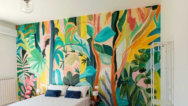 A bedroom with a large tropical-themed mural on the wall, a bed with white bedding, and a bedside table with a lamp.
